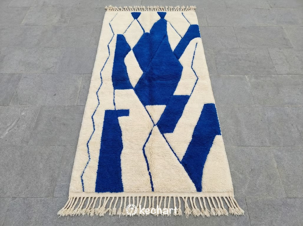 Handwoven custom Beni Ourain Moroccan rug showing plush cream wool texture and vibrant cobalt blue geometric diamond pattern, traditional fringe details.